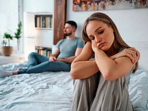Žalostna ženska sedi na postelji, medtem ko je partner v ozadju