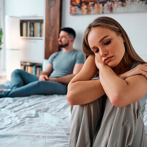 Photograph: couple on bed, woman looking sad in foreground, man turned away