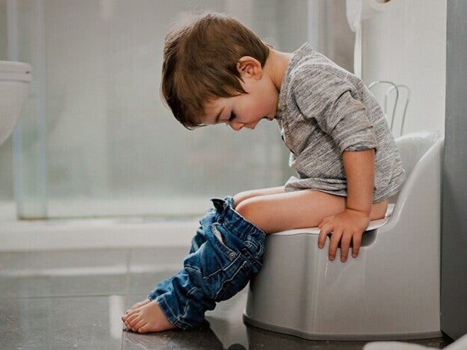 Otrok sedi na kahlici v kopalnici, oblečen v siv pulover in kavbojke.