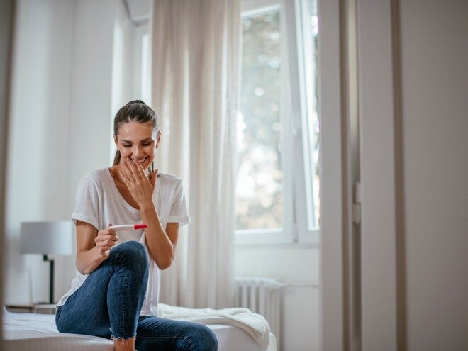 Ženska na postelji z nasmehom gleda na test nosečnosti.