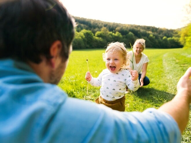 Otrok teče k očetu na travniku, mama sedi v ozadju.