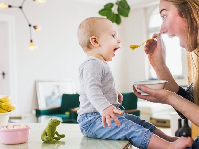 Otrok sedi na mizi in oseba ga hrani z žlico.