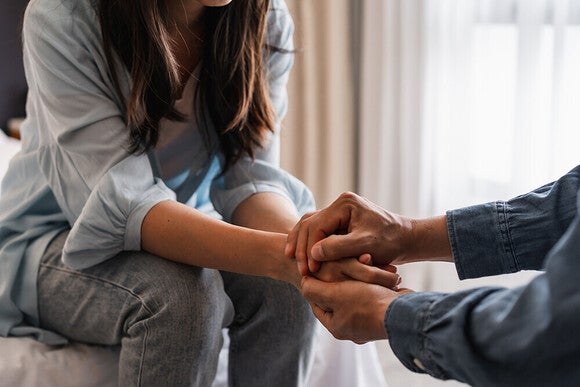 Photo: two people clasping hands in comforting gesture on sofa by bright window