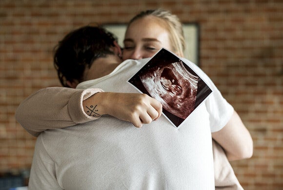 Photograph: smiling woman hugging partner, holding ultrasound photo of fetus