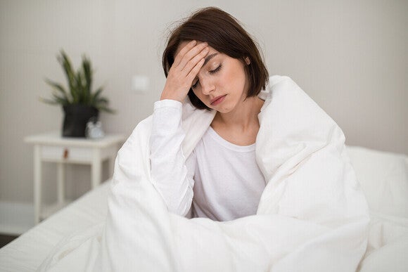Tired woman sitting up in bed, wrapped in white blanket, holding her forehead