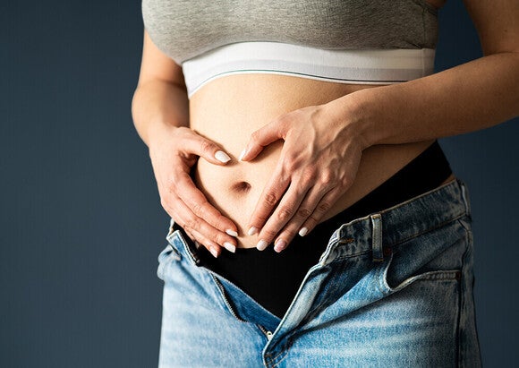 Photograph of pregnant belly with hands forming a heart, gray bra and unbuttoned jeans